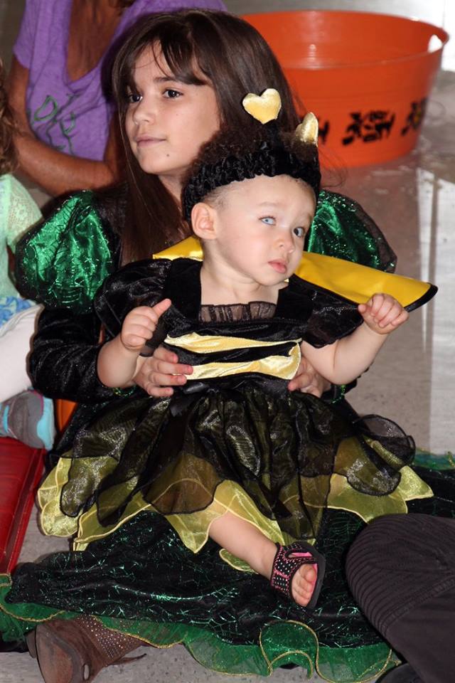 A little girl holding a toddler in her lap. Both children are wearing Halloween costumes.
